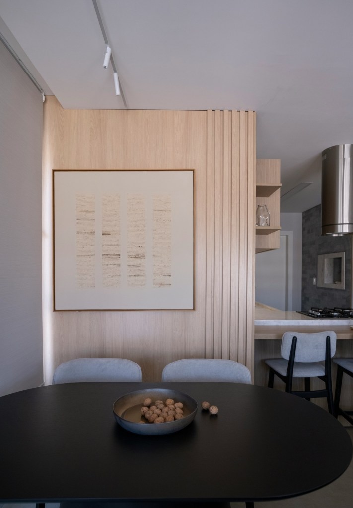 Perete placat cu lemn deschis și lamele verticale, tablou minimalist și masă ovală neagră cu un bol de nuci