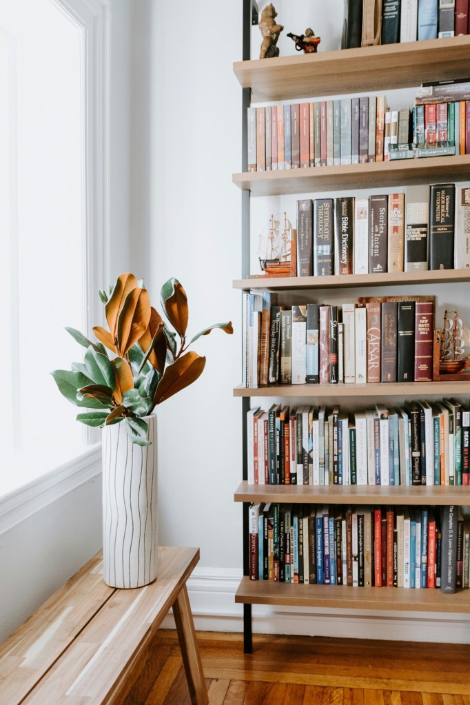 Bibliotecă cu rafturi de lemn deschis, cu cărțile așezate pe teme, lângă o fereastră și o vază cu frunze de magnolie pe o bancă