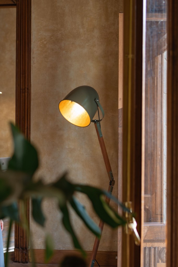 Lampadar verde cu interior auriu lângă un perete de tencuială caldă, între rame de lemn