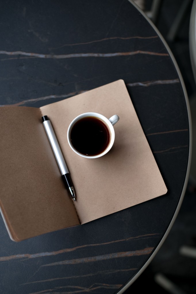Carnet deschis din hârtie kraft, cu un pix și o ceașcă de cafea, pe o masă rotundă închisă la culoare