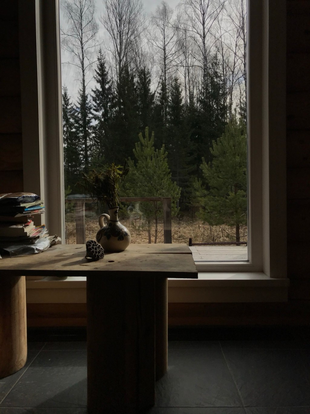 Masă din lemn lângă o fereastră mare cu vedere spre pădure, cu o carafă din ceramică, un fruct uscat și un teanc de reviste