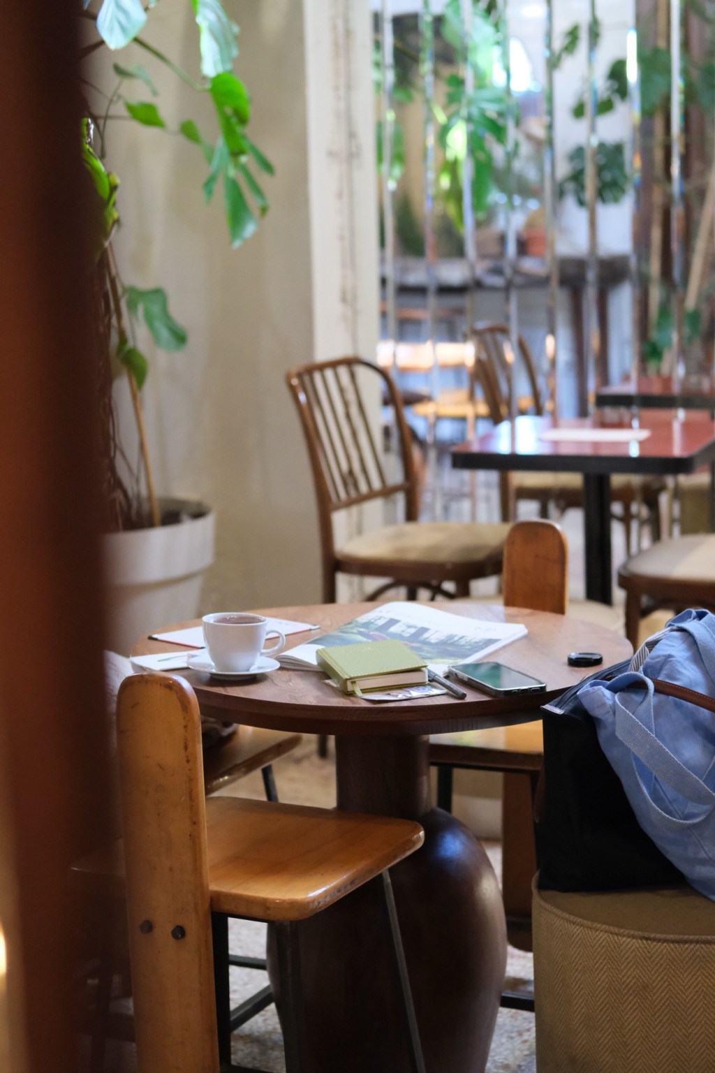 Masă rotundă din lemn cu o ceașcă de ceai, un carnet verde și o revistă de design, într-o cafenea cu plante