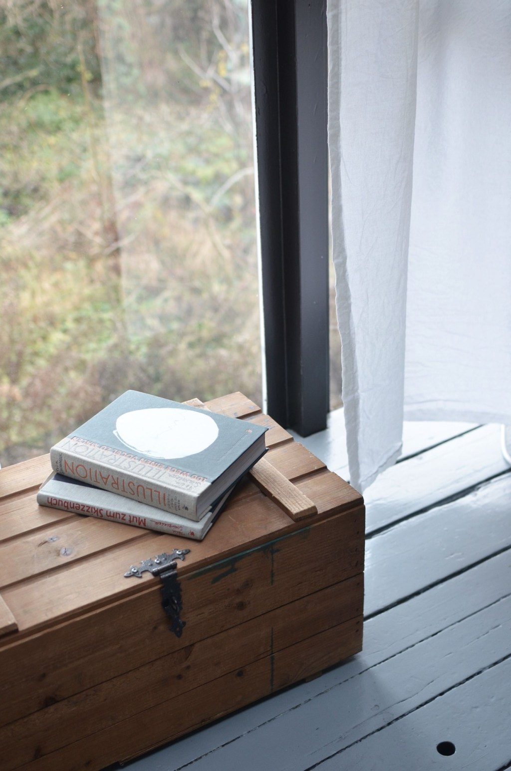 Cufăr vechi de lemn cu două cărți, lângă o fereastră cu perdea albă de in