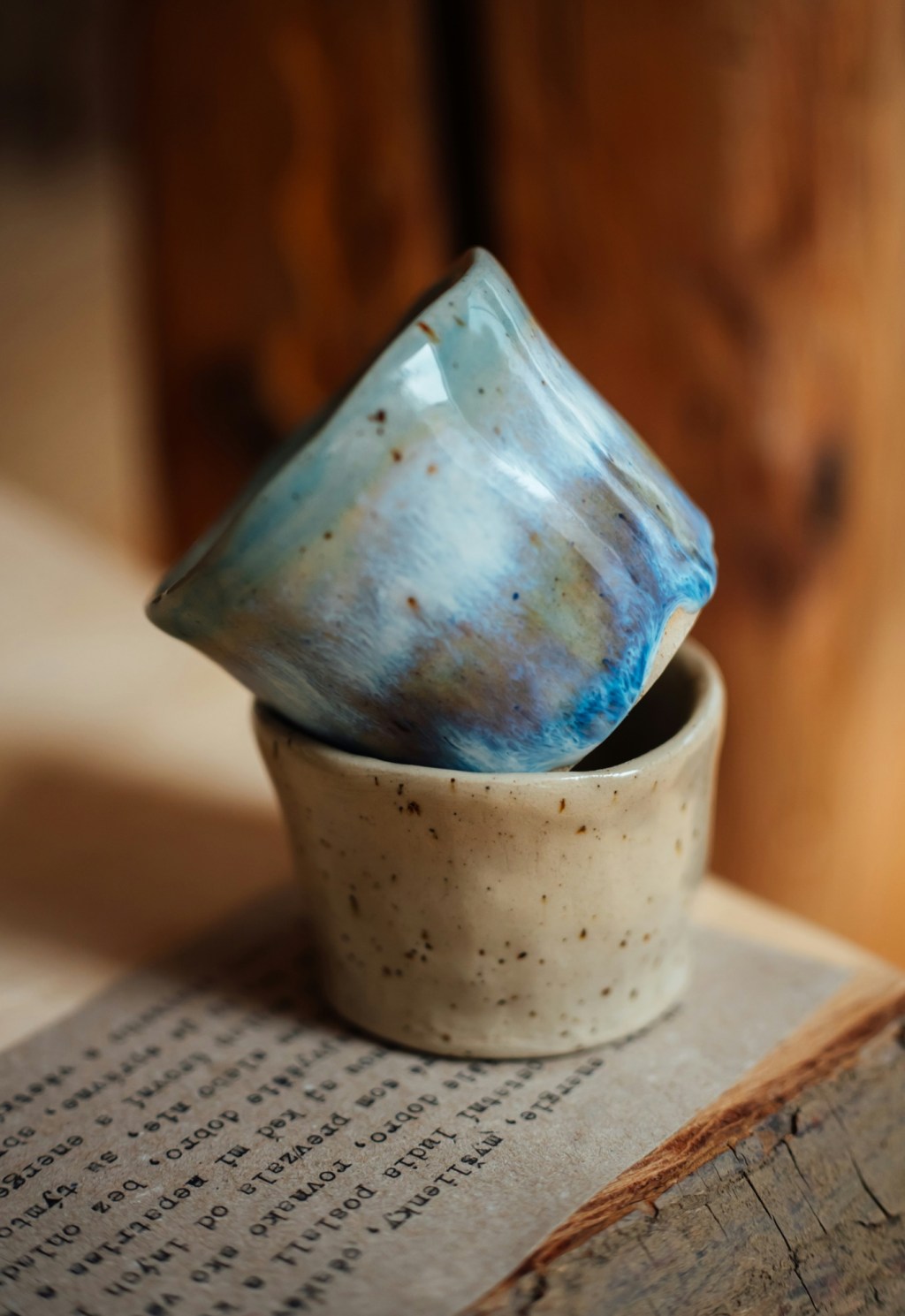 Două căni mici de ceramică lucrate de mână, una cu smalț albastru, cealaltă crem cu pistrui, așezate pe o carte veche